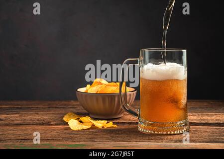Gießen Sie schaumig Bier in einen Becher auf einem Holztisch mit Chips. Oktoberfest. Stockfoto