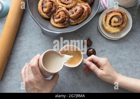Frühstücksszene mit einer weißen Frau, die ein Glas Milch in eine Tasse Kaffee gießt, mit hausgemachten leckeren Zimtschnecken. Stockfoto