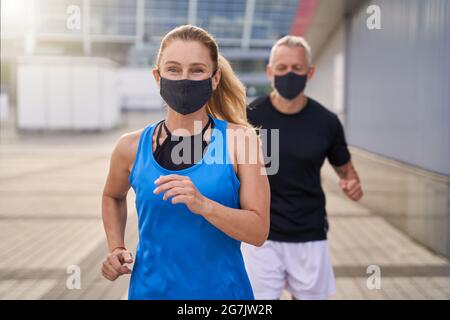 Motivierte, sportliche Frau mit Schutzmaske, während sie mit ihrem Mann läuft, um während der Covid19-Pandemie fit zu bleiben Stockfoto