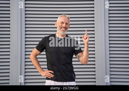 Fröhlicher reifer Mann, der die Kamera anlächelt, die Hand hebt und den Zeigefinger nach oben zeigt, während er draußen auf grauem Hintergrund posiert Stockfoto