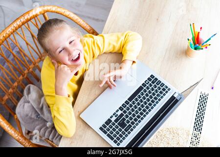 Das kaukasische glückliche Mädchen, 10-11 Jahre alt, sitzt in einem Korbsessel an einem Schreibtisch, neben einem Laptop, zeigt eine Geste - rufen Sie mich an, Draufsicht. Hilfe bei Augenermüdung Stockfoto