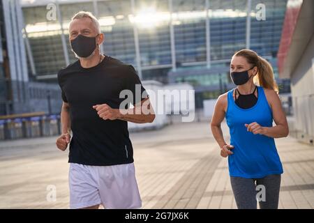 Sportives Paar mittleren Alters joggt morgens mit Schutzmasken. Covid19 Verantwortungsvolles Verhalten Stockfoto