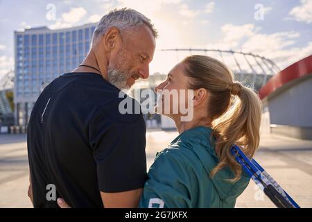 Schönes, sportliches Paar mittleren Alters, das sich gegenseitig anlächelt, während es draußen zusammen steht und sich für das morgendliche Training auf dem Tennisplatz bereit macht Stockfoto