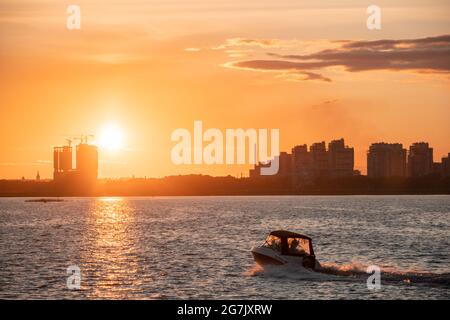 Silhouetten von Stadtgebäuden am Ufer vor dem Hintergrund der untergehenden Sonne und Blendung auf dem Wasser. Sonnenuntergang auf dem Fluss, ein Motorboot crossin Stockfoto