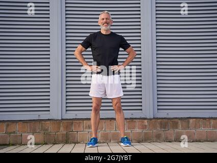 Ganzkörperaufnahme eines aktiven Mannes mittleren Alters in guter Form, der lächelt, während er mit den Armen auf der Taille steht und draußen auf grauem Hintergrund posiert Stockfoto