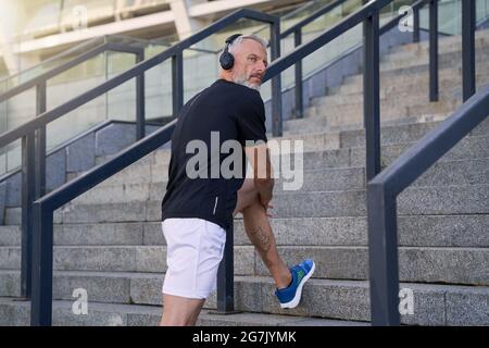 Passen Sie Männer mittleren Alters in Sportkleidung und Kopfhörer, hören Sie Musik oder Podcast und schauen Sie beiseite, während Sie im Freien trainieren Stockfoto