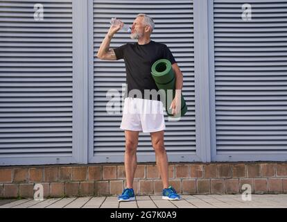 Sportlicher Mann mittleren Alters, der Wasser trinkt, eine Trainingsmatte hält, bereit für Sport im Freien Stockfoto