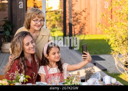 Lächelndes Mädchen, das Selfie mit Großmutter und Mutter am Familientisch im Hinterhof nimmt Stockfoto