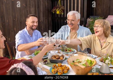 Ein glückliches, aufgeregtes Seniorehepaar, das mit seinen erwachsenen Kindern auf der Familienfeier Gläser mit Alkoholgetränken anklickte Stockfoto