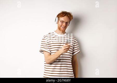 Technologiekonzept. Junger Rotschopf-Hipster in Brille, Musik hören in Kopfbedeckungen, Nachricht lesen auf Smartphone, weißer Hintergrund Stockfoto