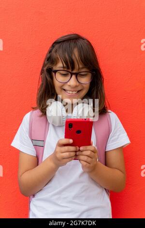Kaukasisches Mädchen mit ihrem Handy und ihren Kopfhörern lächelte vor einer roten Wand sehr. Stockfoto