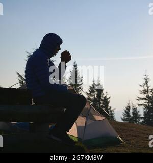 Silhouette eines männlichen Wanderers, der sich nach einer kühlen Nacht im Zelt auf der Bank ausruhte und vor dem Hintergrund einsamer Bäume und eines klaren wolkenlosen blauen Himmels duftenden Kaffee schmeckte. Stockfoto