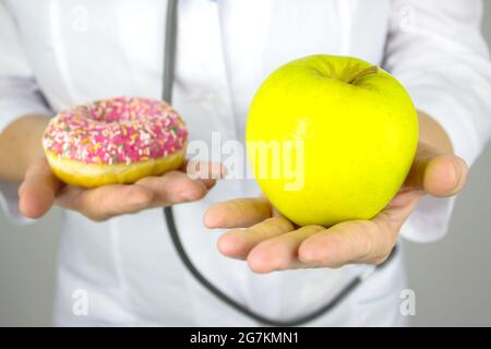 Medizin, Gesundheitskonzept. Nahaufnahme der Hände der Ärztin mit einem Apfel und Donut. Gesunde Ernährung. Das Konzept der Diät Vergleich Stockfoto