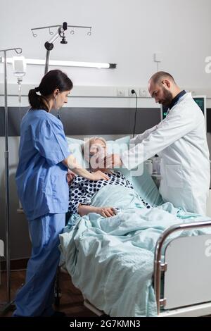 Bewusstlose ältere Patientin, die im Krankenhausbett liegt, und medizinisches Personal hilft ihr mit einer Sauerstoffmaske beim Atmen. Arzt Krankenschwester mit Stethoskop hörende Herz von hospitalisierten älteren Frau. Stockfoto