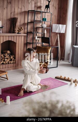 Porträt einer attraktiven, erholsamen Frau in weißen Kleidern, die auf einer hellen Yogamatte mit Kerzen herumsitzt. Gemütliche Holzinterieur mit Kamin mit Baumstämmen. Stockfoto