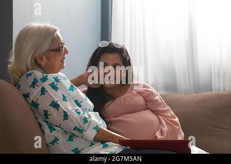 EINE SCHWANGERIN, DIE IHRE MUTTER LIEBEVOLL ANSCHAUT, DIE NEBEN IHR AUF DEM SOFA SITZT Stockfoto