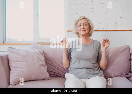 Lächelnde Frau eines älteren Erwachsenen, die im Wohnzimmer Yoga praktiziert. Ältere entspannte weibliche sitzend in Lotushaltung und meditierend Zen wie Stockfoto