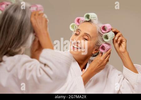 Eine alte Frau stylen Haare mit Rollen vor dem Spiegel. Stockfoto
