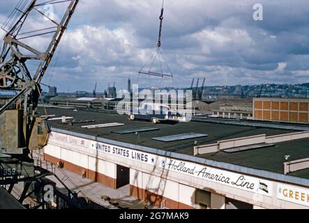 Automobiltransporte per Schiff an den Docks am Hudson River in Hoboken Docks, New Jersey, USA in den späten 1950er Jahren – hier wird auf seiner transatlantischen Reise ein Auto mitten in der Luft ausgesetzt. Die Beschilderung weist darauf hin, dass zu den Lagereinrichtungen an Land die Reedereien Cunard Line, United States Lines und Holland-America Line gehören. Dieses Bild stammt von einem alten Kodak-Amateurfotograf mit Farbtransparenz – einem Vintage-Foto aus den 1950er Jahren. Stockfoto