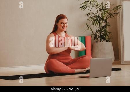 Frau, die Yoga-Meditation macht und online Fitness-Session ansieht. Glückliche Hündin in Sportswear, die zu Hause mit einem Laptop vorne trainiert. Stockfoto