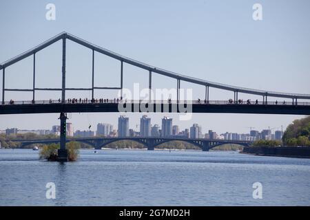 Parkovy Fußgängerbrücke über den Fluss Dnjepr mit Schiffen unter, Kiew, Ukraine. Silhouetten von Menschen, die auf der Brücke spazieren. Brücke bunjee ju Stockfoto
