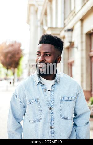 Porträt eines lächelnden jungen afrikanischen Mannes in Jeanshemd, der im Sommer auf der Straße unterwegs ist. Stockfoto