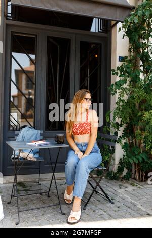 Porträt einer jungen Hipster-Frau in Gläsern, die eine Tasse Kaffee trinkt und ihr Smartphone auf einer Cafeterrasse überprüft, Telearbeit, freiberufliches Konzept. Stockfoto