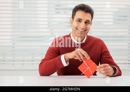 EIN GLÜCKLICHER ERWACHSENER MANN, DER MIT EINEM KLEINEN GESCHENK IM BÜRO SITZT Stockfoto