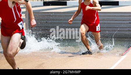 Zwei Läufer, die in roten Uniformen den Hindernislauf bestiegen, landeten und liefen aus dem Wasser und kämpften in einem 3000-Meter-Lauf. Stockfoto