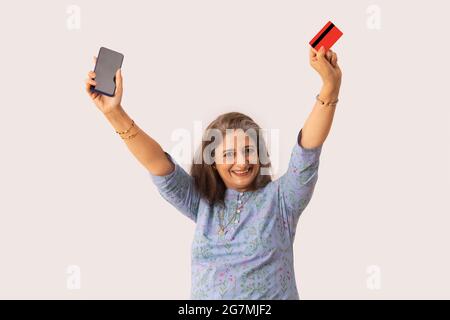 EINE FRÖHLICHE ALTE FRAU, DIE MOBILTELEFON UND DEBITKARTE IN DER HAND HÄLT UND DABEI DIE HÄNDE HEBT Stockfoto