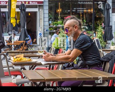 Älterer Mann im Punk-Stil, der Zeitung liest und Chips am Restauranttisch im Café im Freien in Saint Gilles, Brüssel, Belgien, isst. Stockfoto