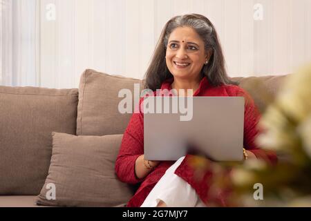 EINE GLÜCKLICHE FRAU, DIE ZU HAUSE SITZT UND WEGSCHAUT, WÄHREND SIE AM LAPTOP ARBEITET Stockfoto