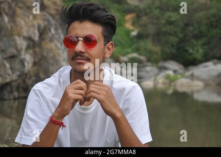 Ein junger Mann aus dem Norden Indiens, der neben dem Fluss sitzt und seitlich schaut, EIN Mann mit weißem T-Shirt und roter Sonnenbrille Stockfoto