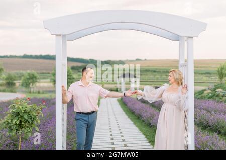 Glücklich zusammen. Schönes, stilvolles Paar mittleren Alters im Lavendelfeld, stehend und sich gegenseitig, lehnend auf dem großen weißen Holzbogen. Nahaufnahme im Hochformat Stockfoto