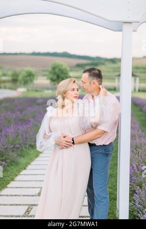 Outdoor-Porträt der schönen fröhlichen romantischen reifen Paar, Mann umarmt Frau, unter dem Holzbogen in Lavendel Feld stehen Stockfoto
