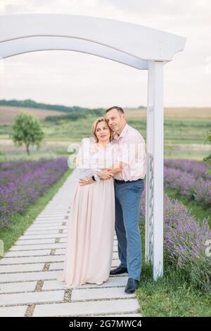 Outdoor-Porträt der schönen fröhlichen romantischen reifen Paar, Mann umarmt Frau, unter dem Holzbogen in Lavendel Feld stehen Stockfoto