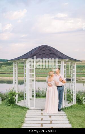 Romantisches Date am Ufer des Teiches. Glückliches reifes Paar, Mann und Frau in eleganten Kleidern, stehen in der Nähe von hölzernen Pavillon am Ufer des schönen Sees Stockfoto