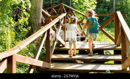 Zwei Mädchen gehen an sonnigen Tagen mit einem Rucksack auf einem Wanderweg zu einem Familiencamp. Schöne Holzstraße im Wald für Touristen. Holztreppe Stockfoto