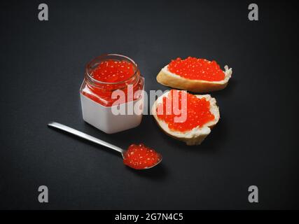 Frischer köstlicher Kaviar. Sandwiches mit rotem Kaviar, Glas und Löffel auf schwarzem Hintergrund. Stockfoto