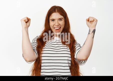 Ja, erfolgreich. Fröhliches Rotschopf-Mädchen, das singt, den Sieg feiert, für das Team wurzelt, die Hände hochhebt, die Faust pumpt und lächelt zufrieden, gewinnt Stockfoto