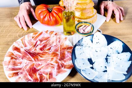 Anonyme Person kocht ein typisch katalanisches oder spanisches Essen. Brot mit Tomate, iberischer Schinkenplatte, Ziegenkäse, Olivenöl und Knoblauch. Traditionelles Essen Stockfoto