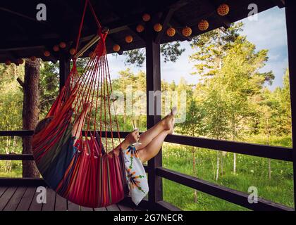 Junge Frau genießt im Sommer die Zeit allein im Waldresort im nordischen Pinienwald, entspannen Sie sich im Schaukel hängenden Textilstuhl auf dem Balkon, trinken Stockfoto