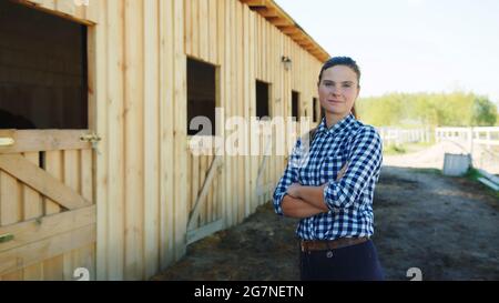 Mädchen, das tagsüber vor dem Pferdestall mit Reihen von Stallfenstern stand. Weibliche Reiterin posiert für die Kamera mit gekreuzten Armen. Blick auf den Pferdestall. Stockfoto
