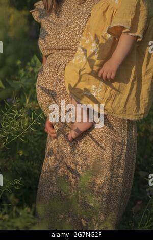 Eine Schwangerin in einem cremefarbenen Kleid hält ihre Tochter auf einem Feld Stockfoto