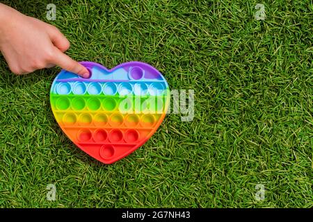Ein Kind spielt mit einem bunten Regenbogen Anti-Stress Sinnesspielzeug Pop es in Form eines Herzens auf dem Gras. Kinderhand platzt Kugeln auf ein Spielzeug Pop it in Stockfoto