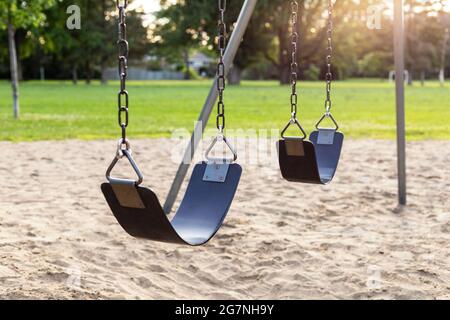 Spielplatz ohne Kinder im öffentlichen Park, Schaukeln auf Ketten mit Sand darunter, Gras, Bäume und Fußballplatz im Hintergrund Stockfoto