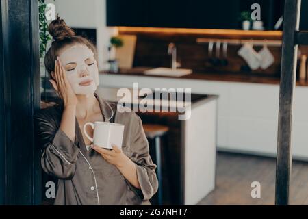 Junge Frau im Seiden-Pyjama, die zu Hause bleibt und morgens eine Tasse Kaffee genießt Stockfoto