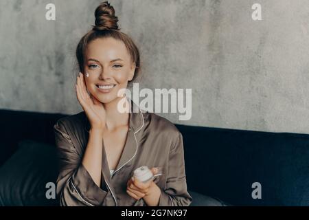 Bild einer fröhlichen kaukasischen Frau im Seidenpyjama, die auf dem Bett sitzt Stockfoto