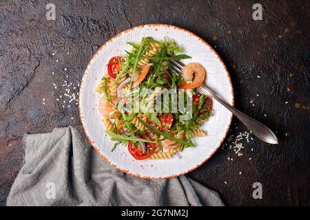 Draufsicht auf leckere Pasta mit Garnelen, Kirschtomaten, Zwiebeln, Sesam und grünen Rucola-Blättern auf dunklem Beton-Hintergrund Stockfoto