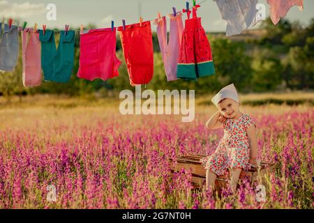 Kleines süßes Baby, das auf einer Holzkiste im Lavendelfeld sitzt und für die Kamera posiert. Bunte Kleidung hängt am Seil auf dem Feld Stockfoto